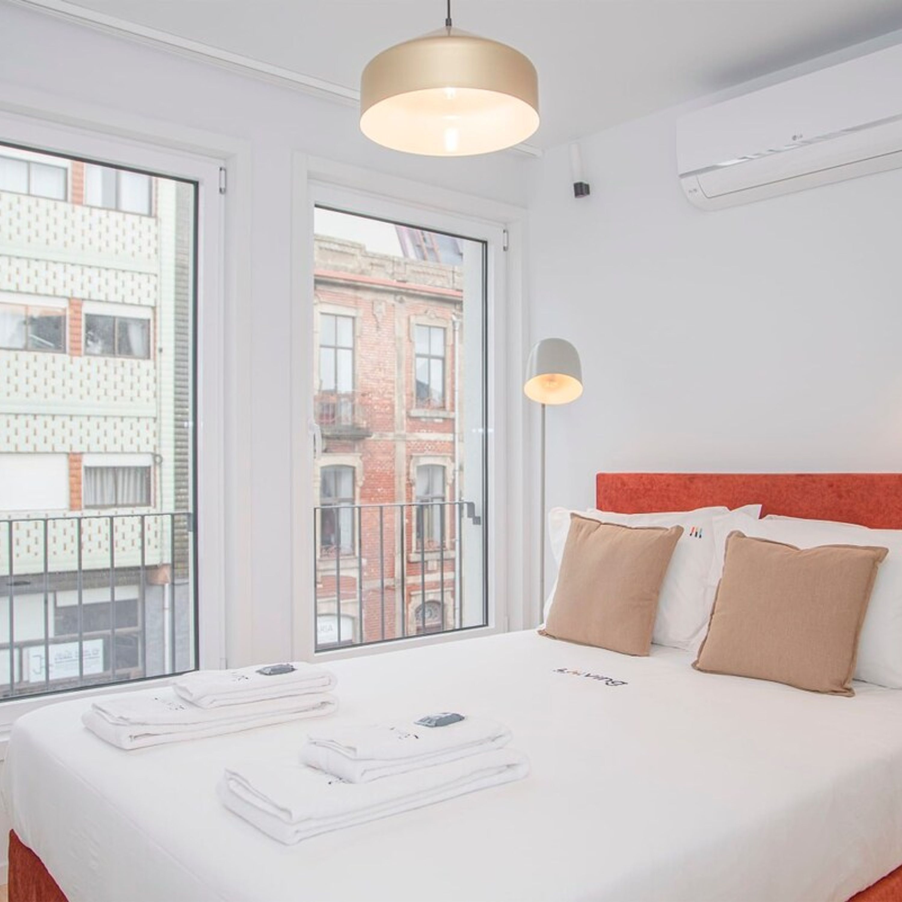 A hotel room with a large bed, a red headboard, and a view of a cityscape through the window.