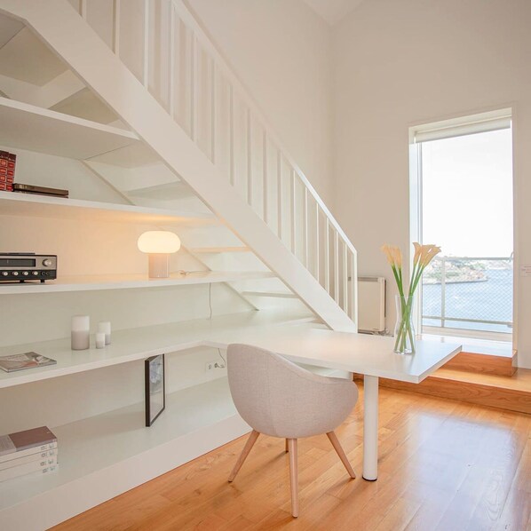 A modern, minimalist room with a white staircase, a desk with a chair, and a view of the sea through a large window.
