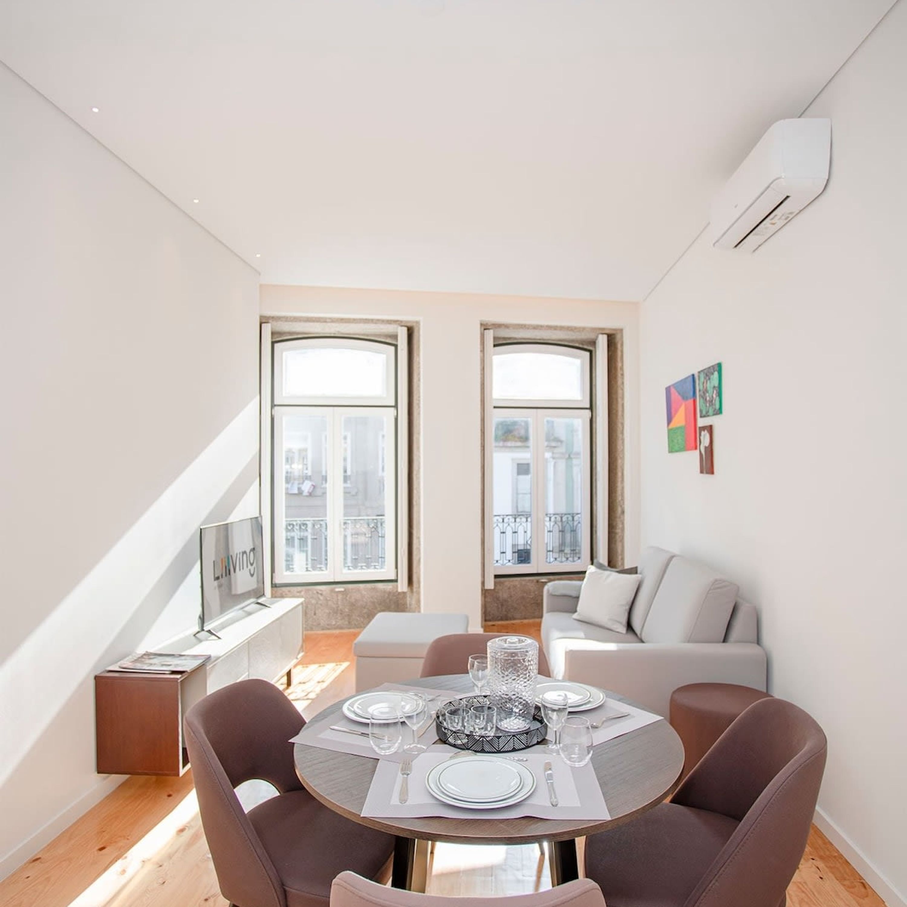 Un comedor moderno con una mesa redonda preparada para cuatro comensales, dos ventanas que aportan luz natural, un sofá gris y una pequeña mesita con un cuadro enmarcado.