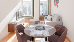 A modern dining area with a round table set for four, two windows providing natural light, a gray sofa, and a small side table with a framed picture. - Liiiving in Porto-mouzinho Residence Apartment 2F (Porto)