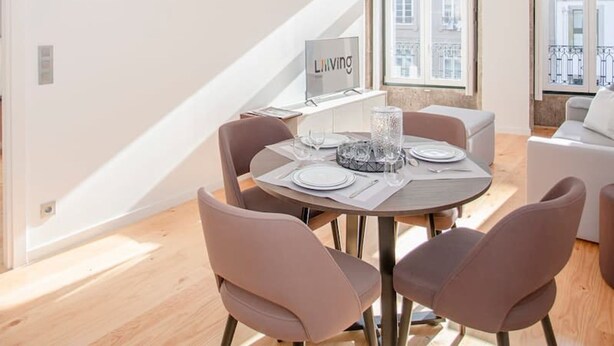 A modern dining area with a round table set for four, surrounded by cushioned chairs. A large window provides a view of the cityscape. - Liiiving in Porto-mouzinho Residence Apartment 2F (Porto)