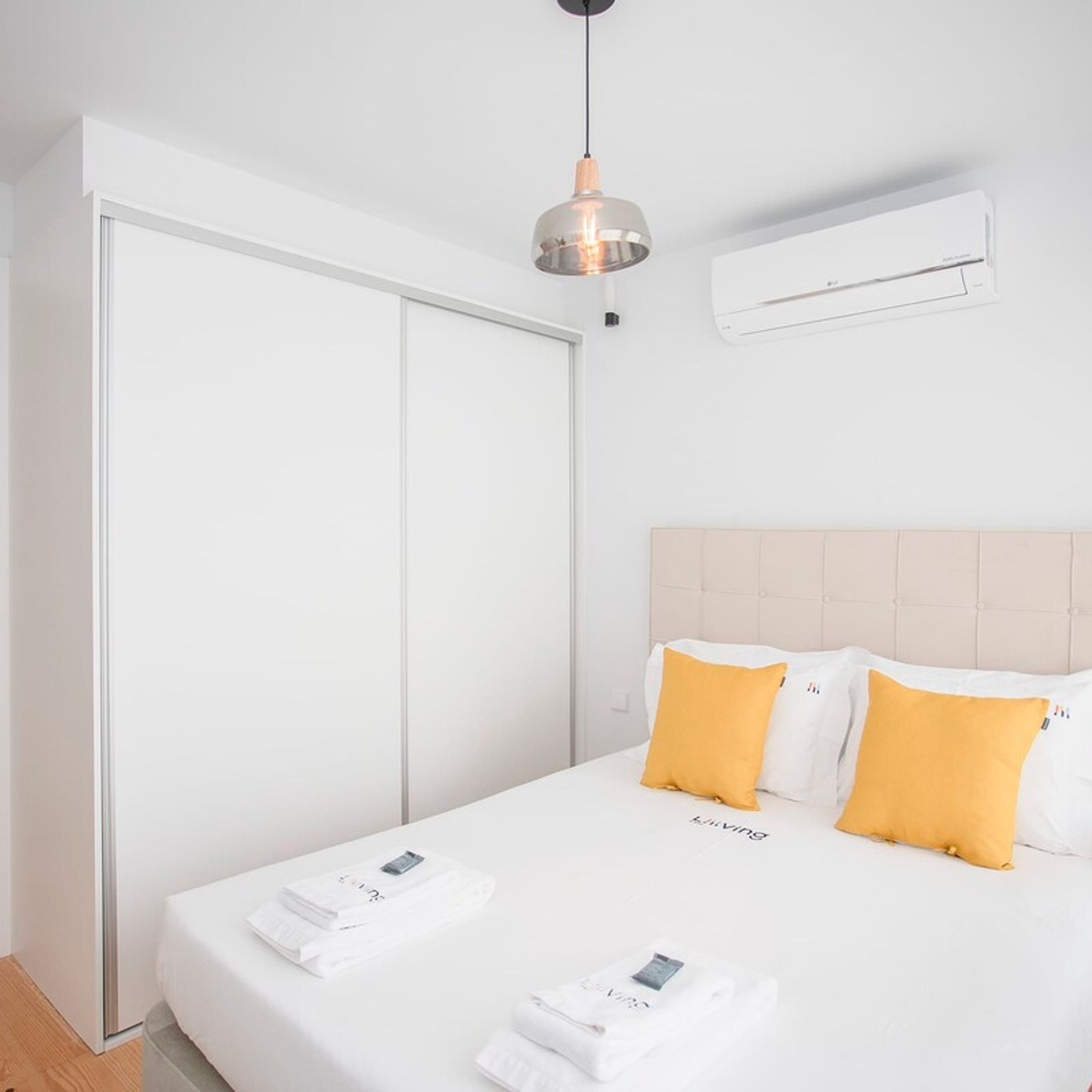 Une chambre d’hôtel avec un lit, du linge de lit blanc, deux oreillers jaunes, un luminaire suspendu et un appareil de climatisation fixé au mur.