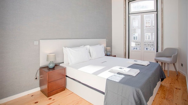 A modern bedroom with a large bed, a nightstand, a chair, and a window with a view of buildings. - Liiiving in Porto-mouzinho Residence Apartment 3F (Porto)