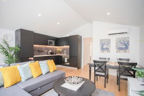 Un salon moderne avec un canapé gris, des coussins jaunes, une table basse ronde et un coin repas comprenant une table et des chaises en bois foncé.