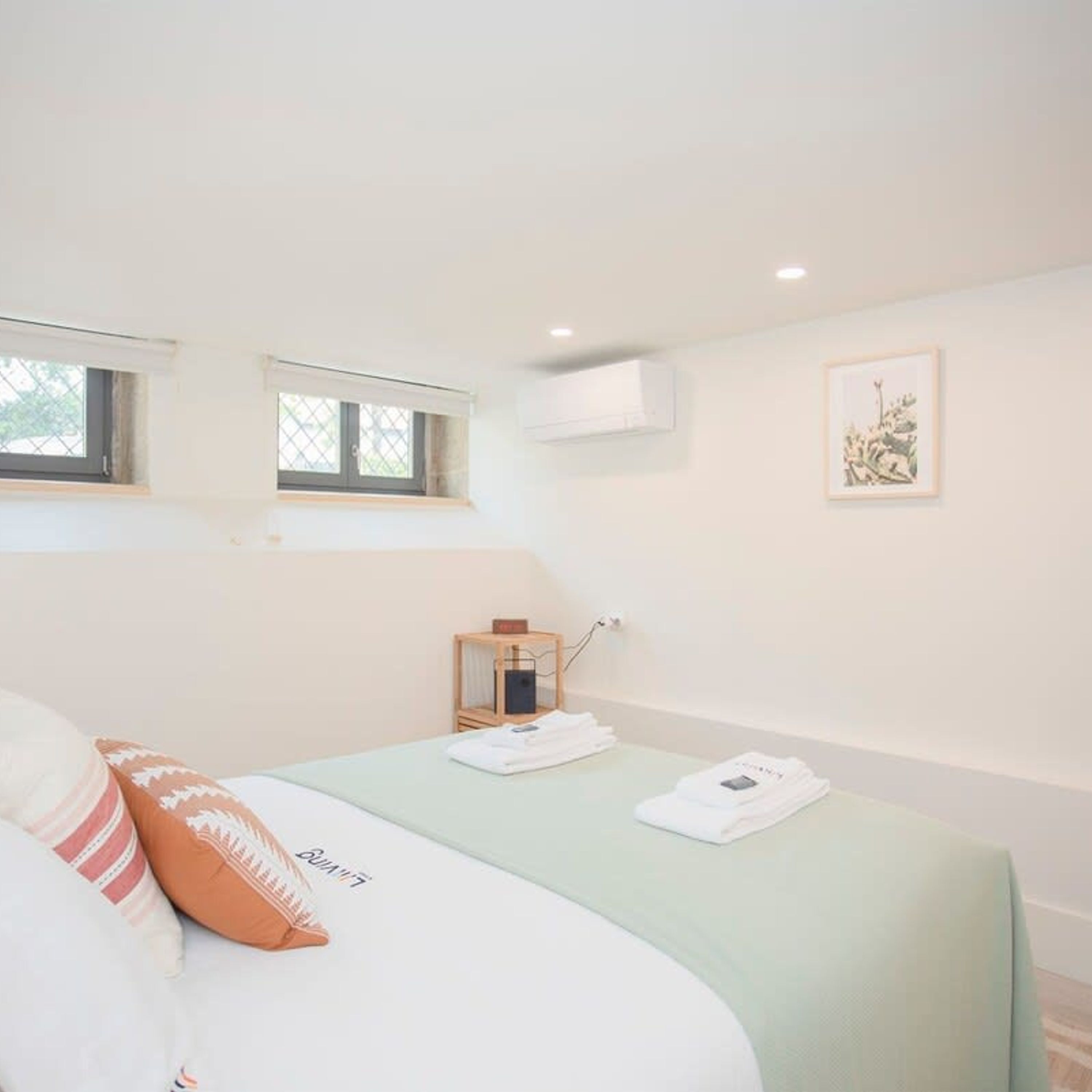 Een slaapkamer met een bed, kussens, een nachtkastje met een lamp, een raam en een lijst met een afbeelding aan de muur.