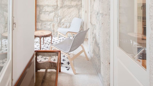A narrow corridor with a stone wall, wooden ceiling, and a glass door leading to a room. There is a wicker chair and a small round table with a woven top. - Liiiving in Porto - Oasis Retreat Apartment (Porto)