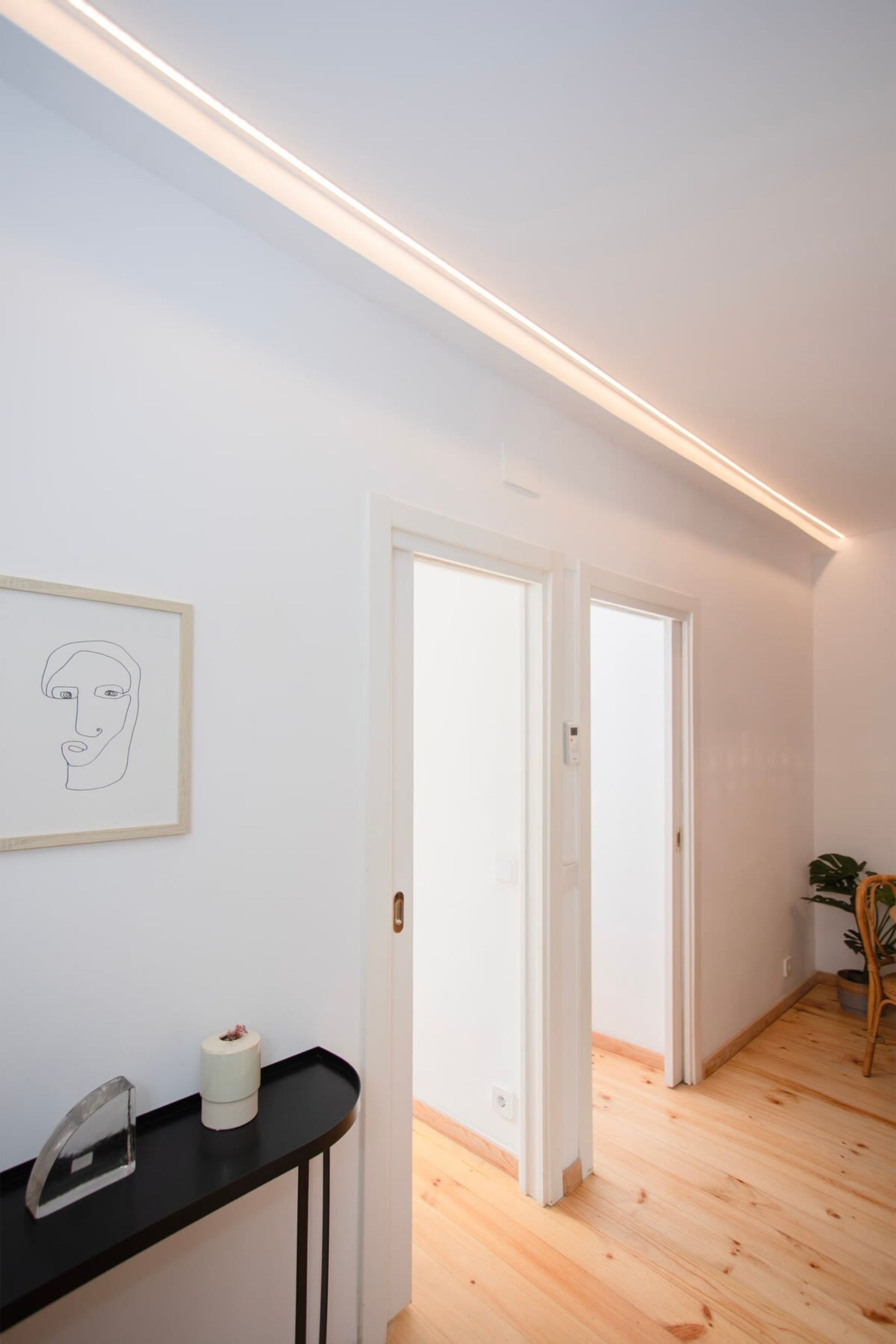 A hallway with a wooden floor, white walls, and a framed artwork. There is a black console table with a clear glass object and a white cylindrical item. Two white doors are visible, one open, leading to another room.