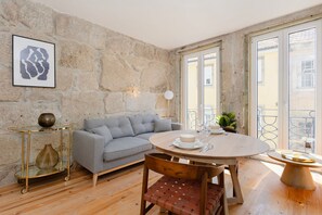 Un salon chaleureux avec un mur en pierre, un canapé gris, une table à manger ronde en bois et une petite table d’appoint sur laquelle est posé un vase.