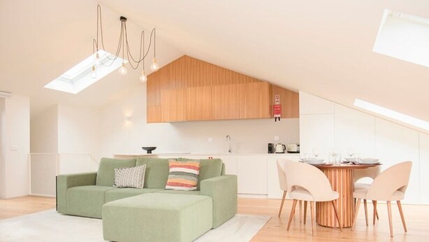 A modern loft apartment with a high ceiling, a dining area with a wooden table and chairs, a green sofa set, and a kitchen with wooden cabinets. - Liiiving in Porto - Cedofeita Green Terrace (Porto)