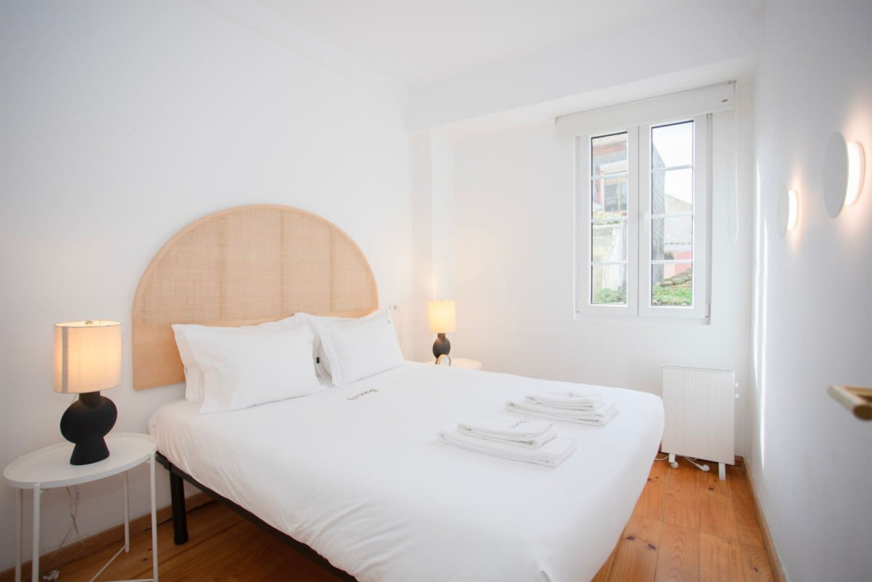 A hotel room with a large bed, two bedside lamps, a window with blinds, and a radiator.