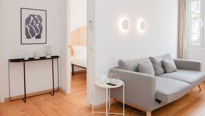 A modern, well-lit room with a grey sofa, a small white side table, a framed abstract artwork, and a bed in the background. - Liiiving in Gaia - Casa da Vinha Apartment V (Vila Nova de Gaia)