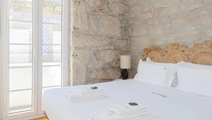 A hotel room with a stone wall, a bed with white linens, a bedside lamp, and a view through a glass door. - Liiiving in Gaia - Casa da Vinha Apartment VI (Vila Nova de Gaia)