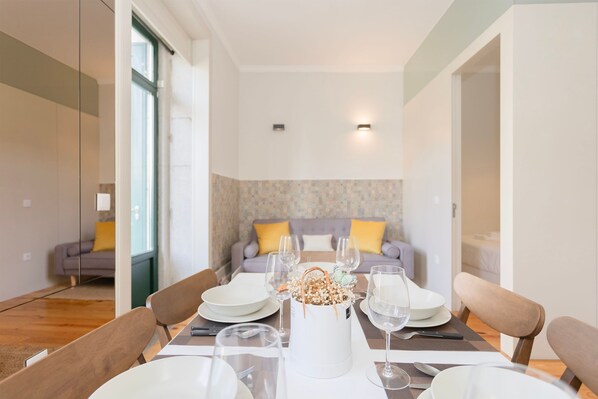 Uma sala de jantar com uma mesa posta para quatro pessoas, um sofá ao fundo e um arranjo decorativo.