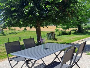 A metal table with four chairs, a vase with purple flowers, and a tree in the background. - Charming Countryside Home with Modern Amenities in Scenic Perche Hills (Melleray)
