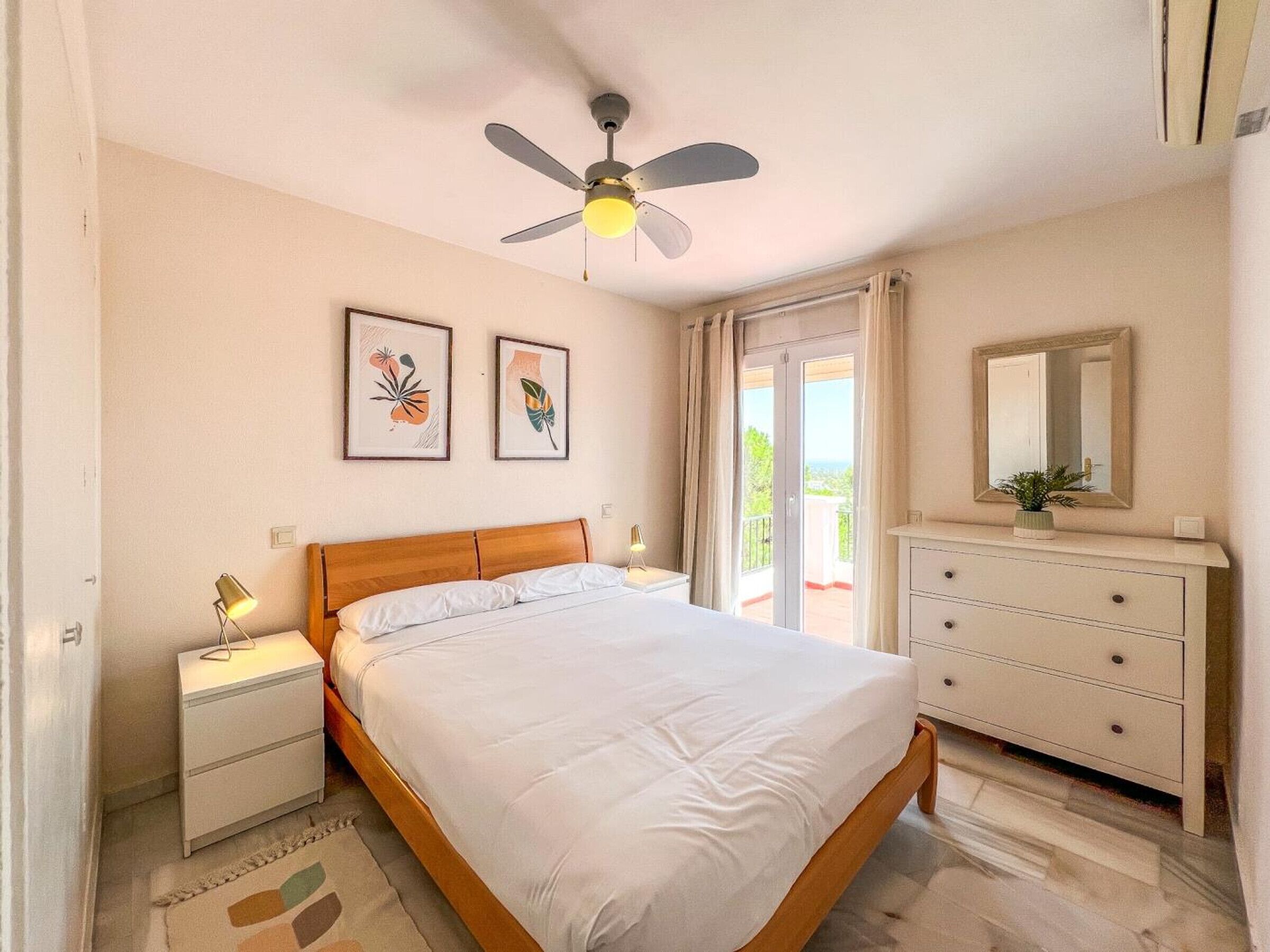 Une chambre à coucher comprenant un lit, une table de chevet, une commode, un ventilateur de plafond et deux œuvres d’art encadrées accrochées au mur.