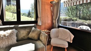 A cozy room with a wooden interior, a sofa, a chair, and large windows offering a view of greenery and a building. - Renovated T2 in Le Grand-Bornand with balcony, parking and terrace, 4 people, near village (Le Grand-Bornand)