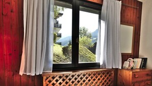 A bedroom with a wooden headboard, a window with white curtains, and a view of greenery and a building outside. - Renovated T2 in Le Grand-Bornand with balcony, parking and terrace, 4 people, near village (Le Grand-Bornand)