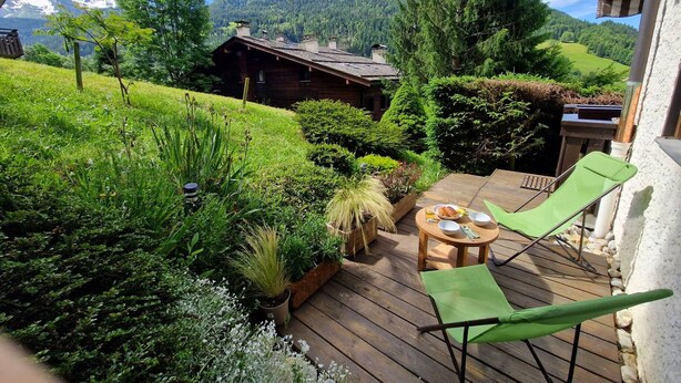 A wooden deck with a table and chairs, surrounded by lush greenery and a mountain backdrop. - Renovated T2 in Le Grand-Bornand with balcony, parking and terrace, 4 people, near village (Le Grand-Bornand)