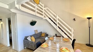 A dining area with a wooden table set for four, a grey sofa with cushions, a white staircase, and a hanging basket. - 3-room apartment with pool, AC and Wi-Fi in Porticcio (Porticcio)
