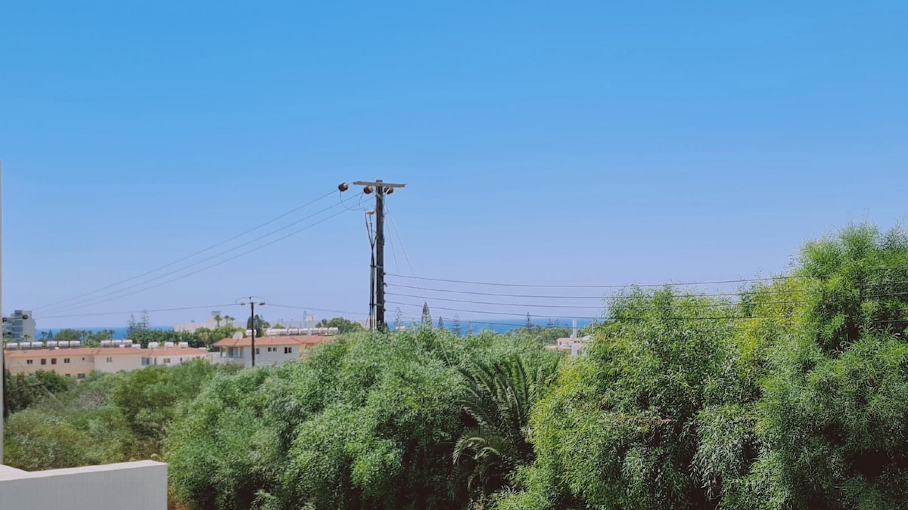 Zona residencial con un alto poste de electricidad y líneas de tendido, rodeada de abundante vegetación y edificios al fondo.
