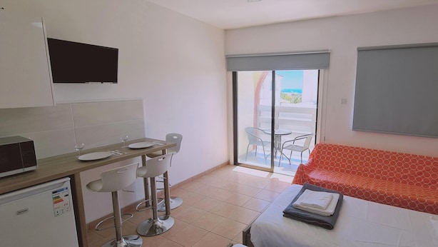 Una habitación compacta con kitchenette, un comedor con mesa y sillas, un sofá y una puerta corrediza de vidrio que da a un espacio exterior.