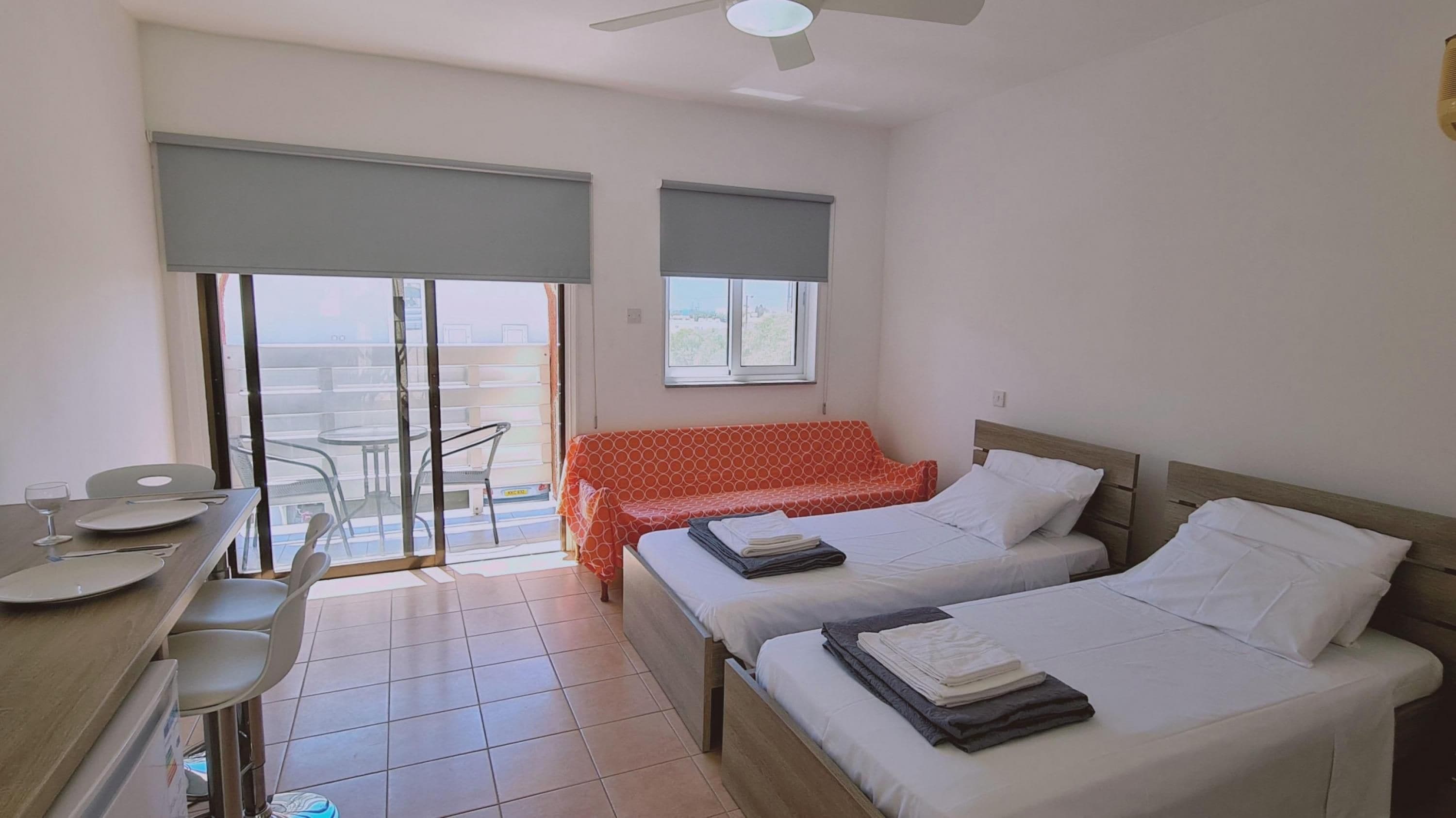 Camera d'albergo con due letti, un tavolo da pranzo con sedie, un balcone con tavolo e sedie e una ventola a soffitto.