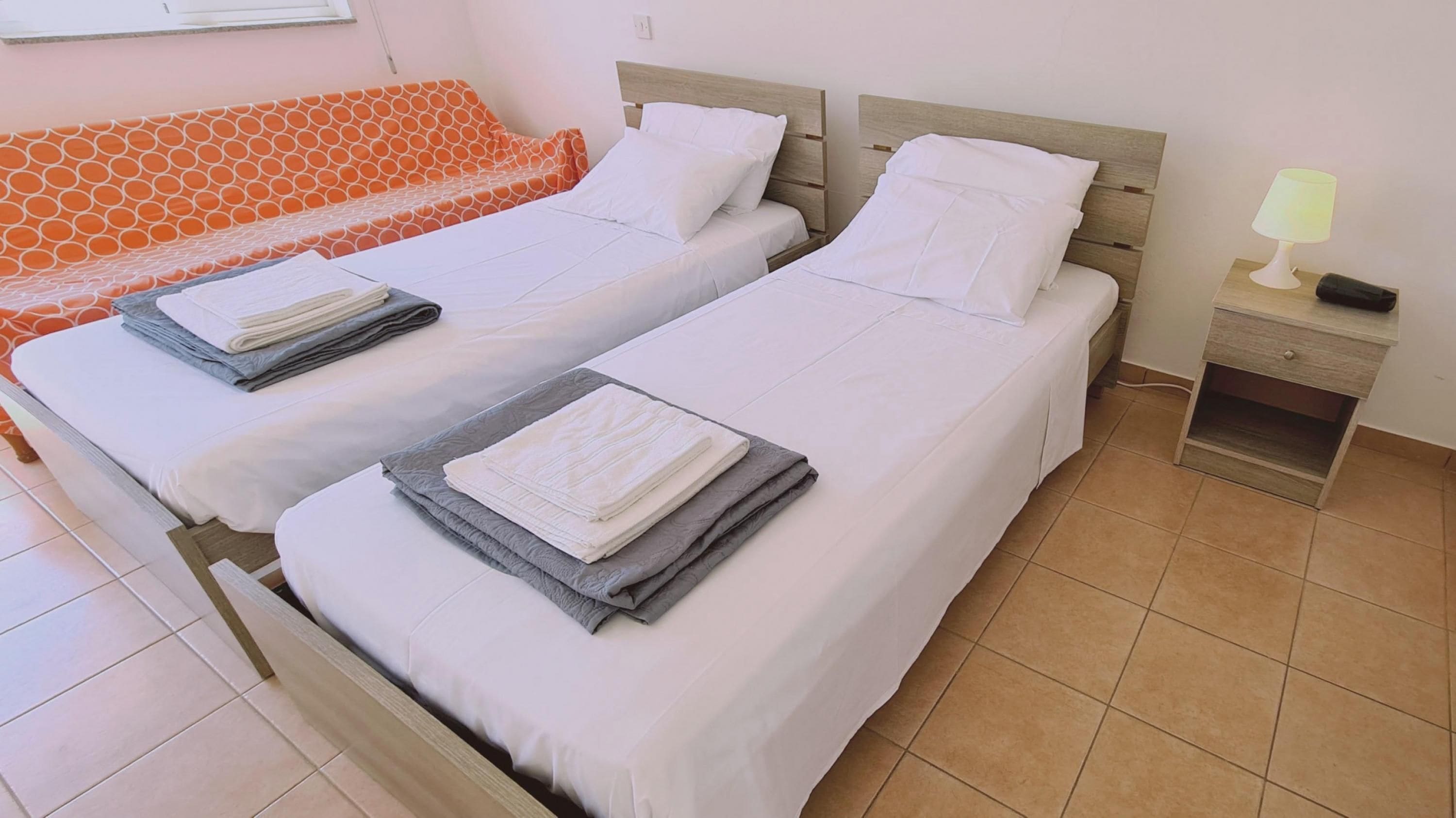 Une chambre d’hôtel avec deux lits simples, un canapé, une table de chevet avec une lampe et un sol carrelé.