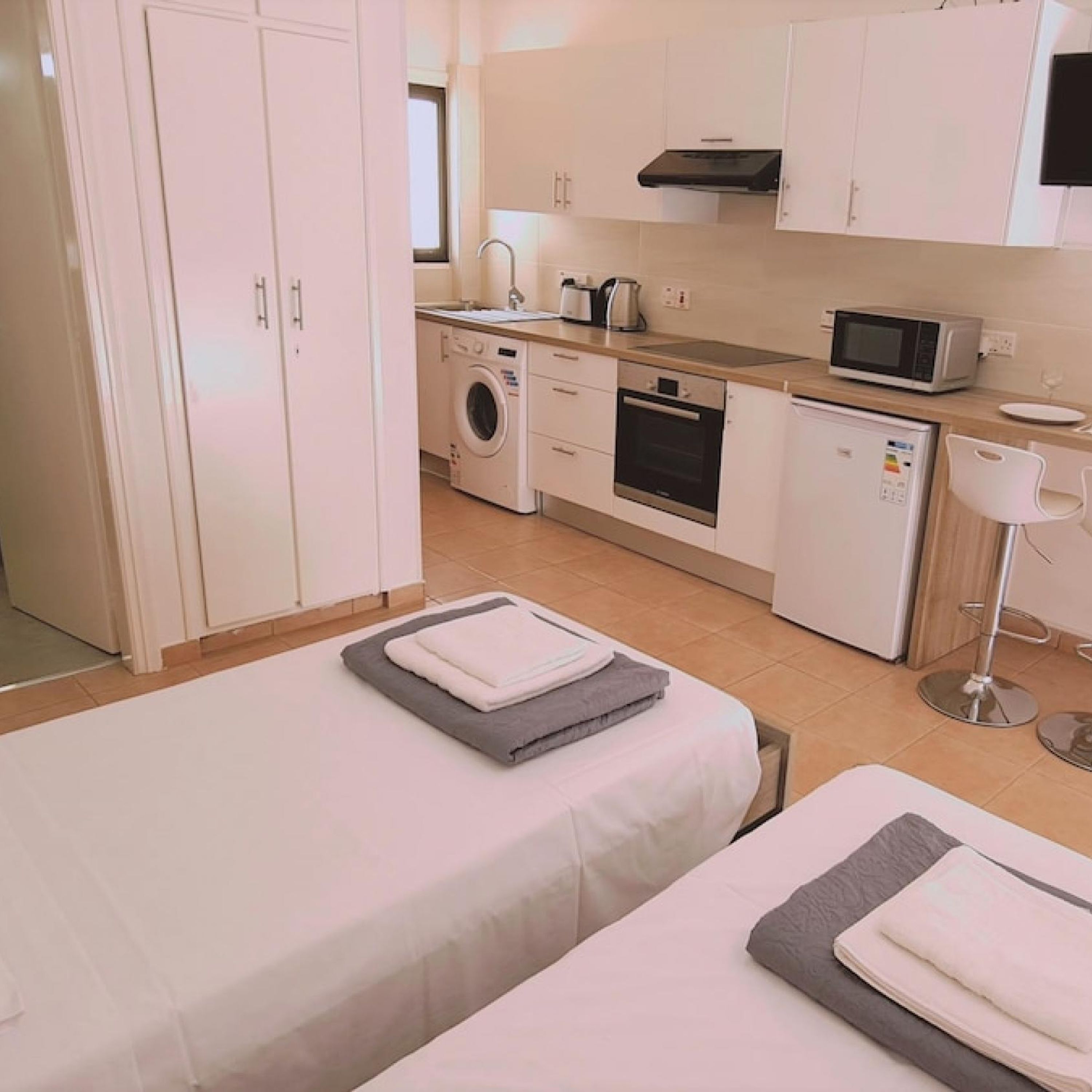 A compact hotel room with two beds, a washing machine, an oven, a microwave, and a small dining area.