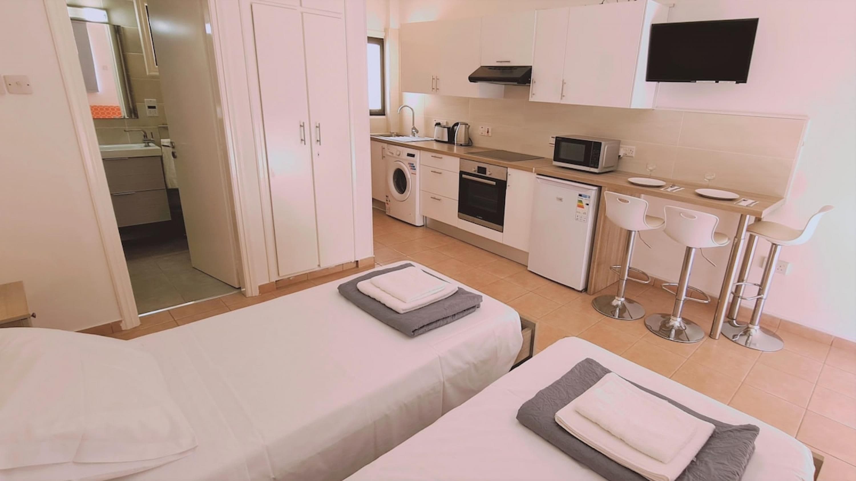 Camera d'albergo compatta con due letti singoli, angolo cottura con microonde, lavatrice e piccolo spazio pranzo con sgabelli da bar.