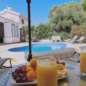 A poolside table with fruit and drinks, a pool, and a building with a white facade and shutters. - 15 ATH - Swallow s Nest Villa (Sotira)
