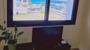 A room with a large window overlooking a coastal area, a television on a stand, and a potted plant on a side table. - 20 ATR -trinity 1 Apartment (Paralimni)