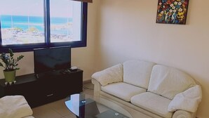 A living room with a sofa, a coffee table, a TV, and a window with a view of the beach. - 20 ATR -trinity 1 Apartment (Paralimni)