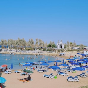 Une plage avec de nombreux transats et parasols, un ciel bleu dégagé et quelques personnes qui se baignent.