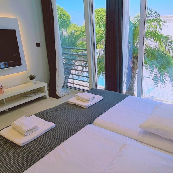 A hotel room with two beds, a TV, a small table with a plant, and a view of palm trees and a pool. - 116 AN 2 - Mezarina 2 Apartment (Ayia Napa)
