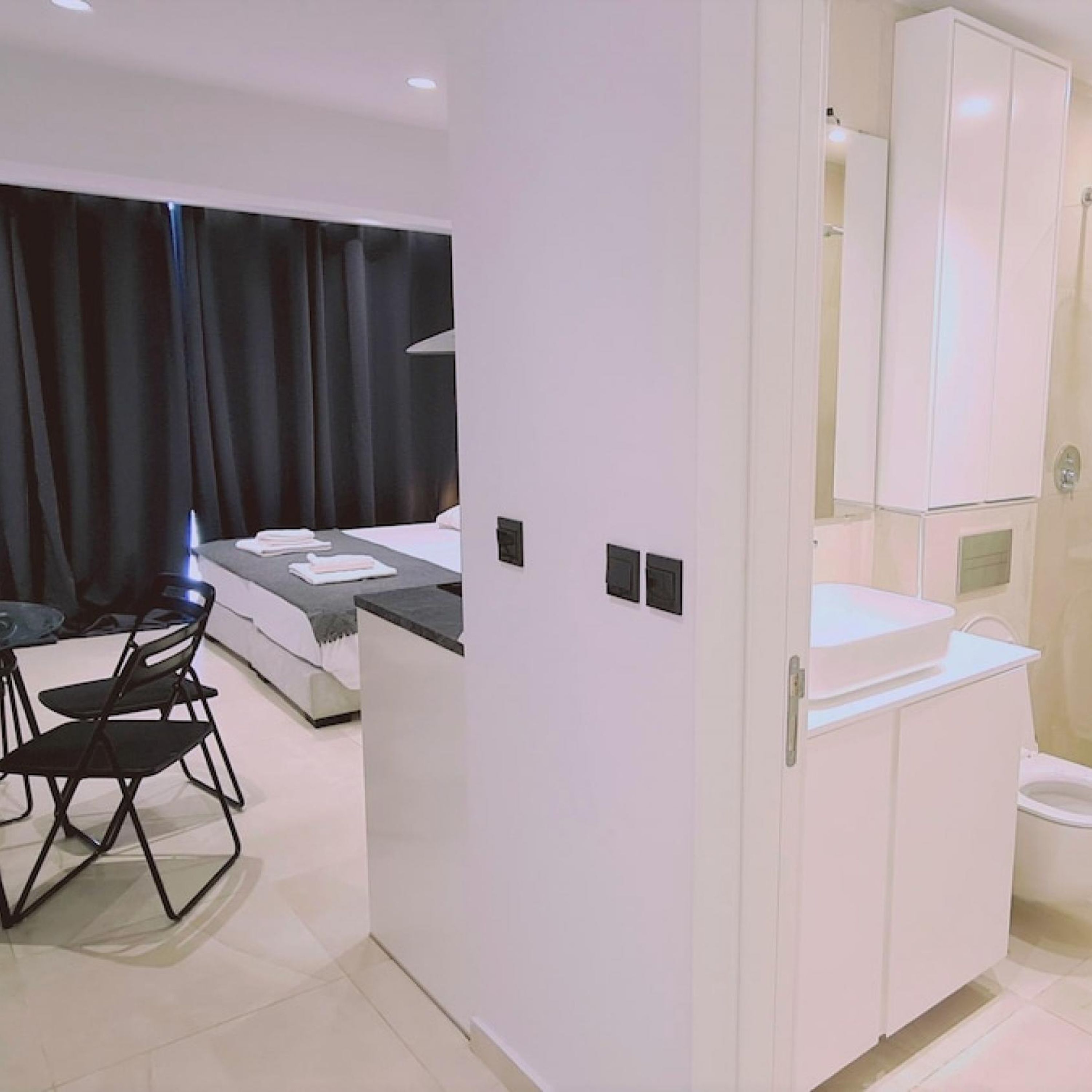 Una habitación de hotel moderna con una cama, una pequeña mesa con sillas, un baño con lavamanos y espejo, y un amplio ventanal con cortinas.