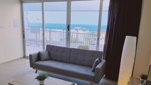 A modern living room with a grey sofa, a wooden coffee table, and a view of the sea through large windows. - 40 PRO 2 - Sunrise Beach Apt 2 (Paralimni)