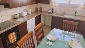 A compact kitchen with dark wooden cabinets, a granite countertop, a built-in oven, and a dining area with a glass table set for two. - 40 PRO 2 - Sunrise Beach Apt 2 (Paralimni)