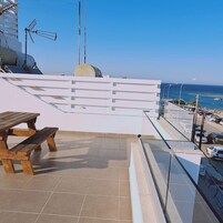 Una terraza en la azotea con una mesa y sillas de madera, barandal blanco y vista despejada al mar y coches estacionados abajo.