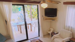 Un salon comprenant un canapé, une table basse, un téléviseur fixé au mur et une porte-fenêtre coulissante donnant sur un espace extérieur avec une table et des chaises.