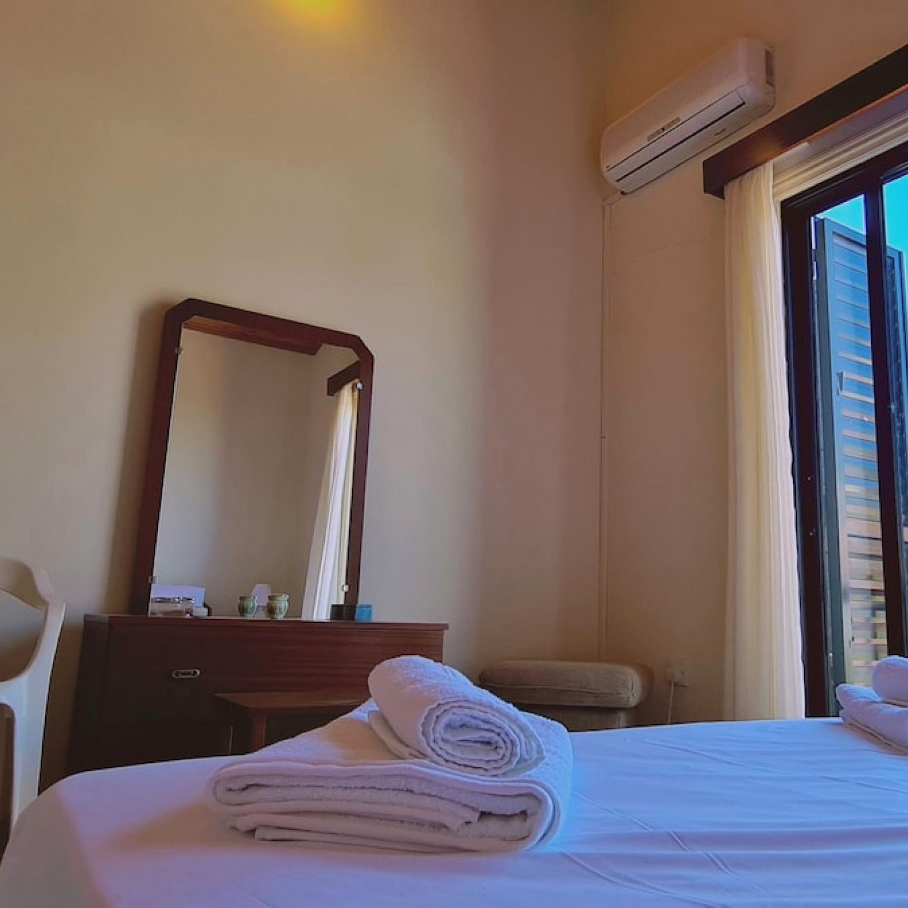 A hotel room with a bed, a dresser, a mirror, an air conditioning unit, and a window with curtains.