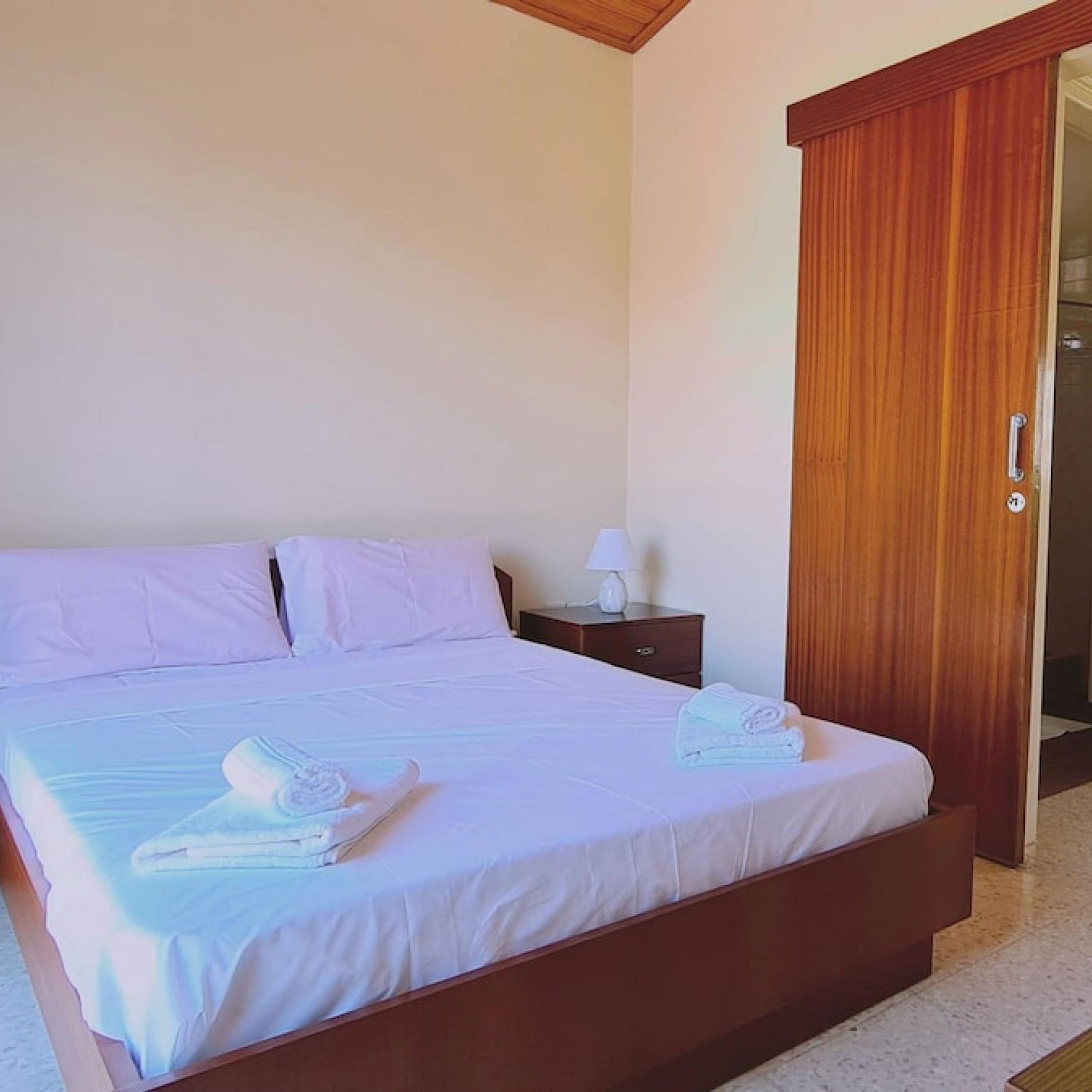 A bedroom with a bed, pillows, a nightstand, a lamp, and a wooden wardrobe.
