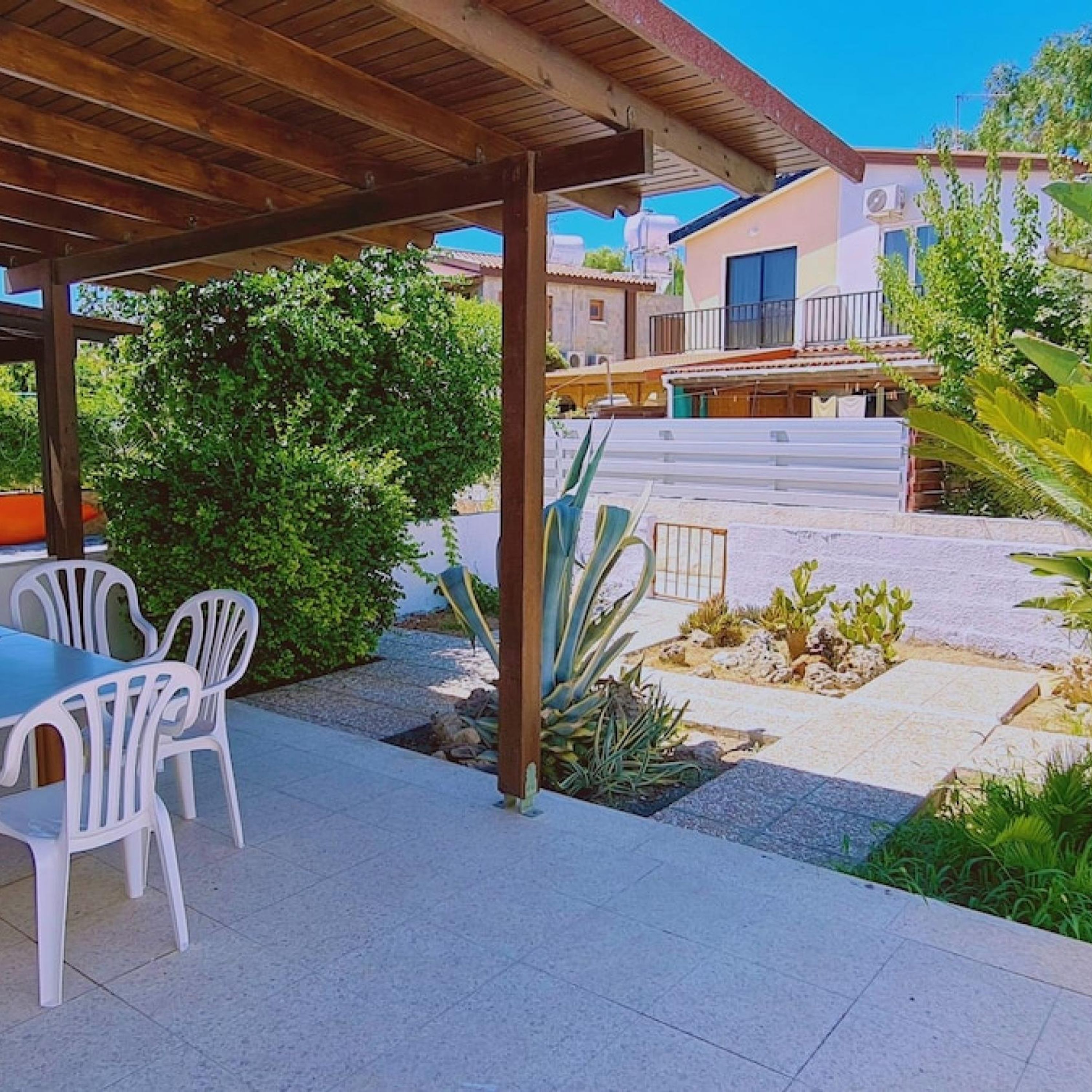 An outdoor patio area with a wooden pergola, white plastic chairs, and a variety of plants including an agave.