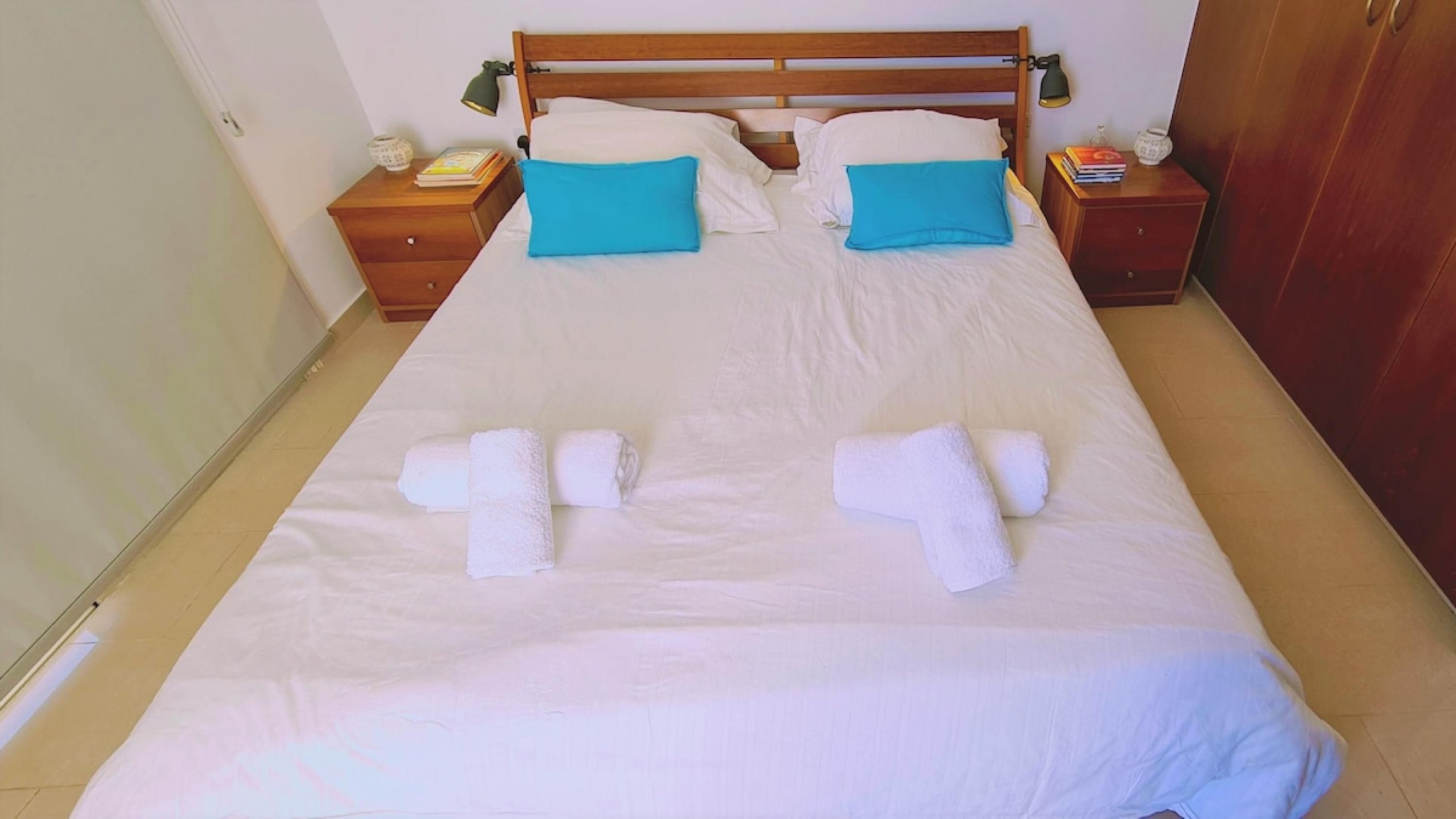 A neatly made bed with white linens and two blue pillows, a wooden headboard, and two bedside tables with lamps and books.