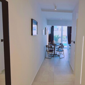 Un estrecho pasillo que conduce a un comedor con una mesa y sillas, un cuadro en la pared y vistas al exterior a través de un ventanal.