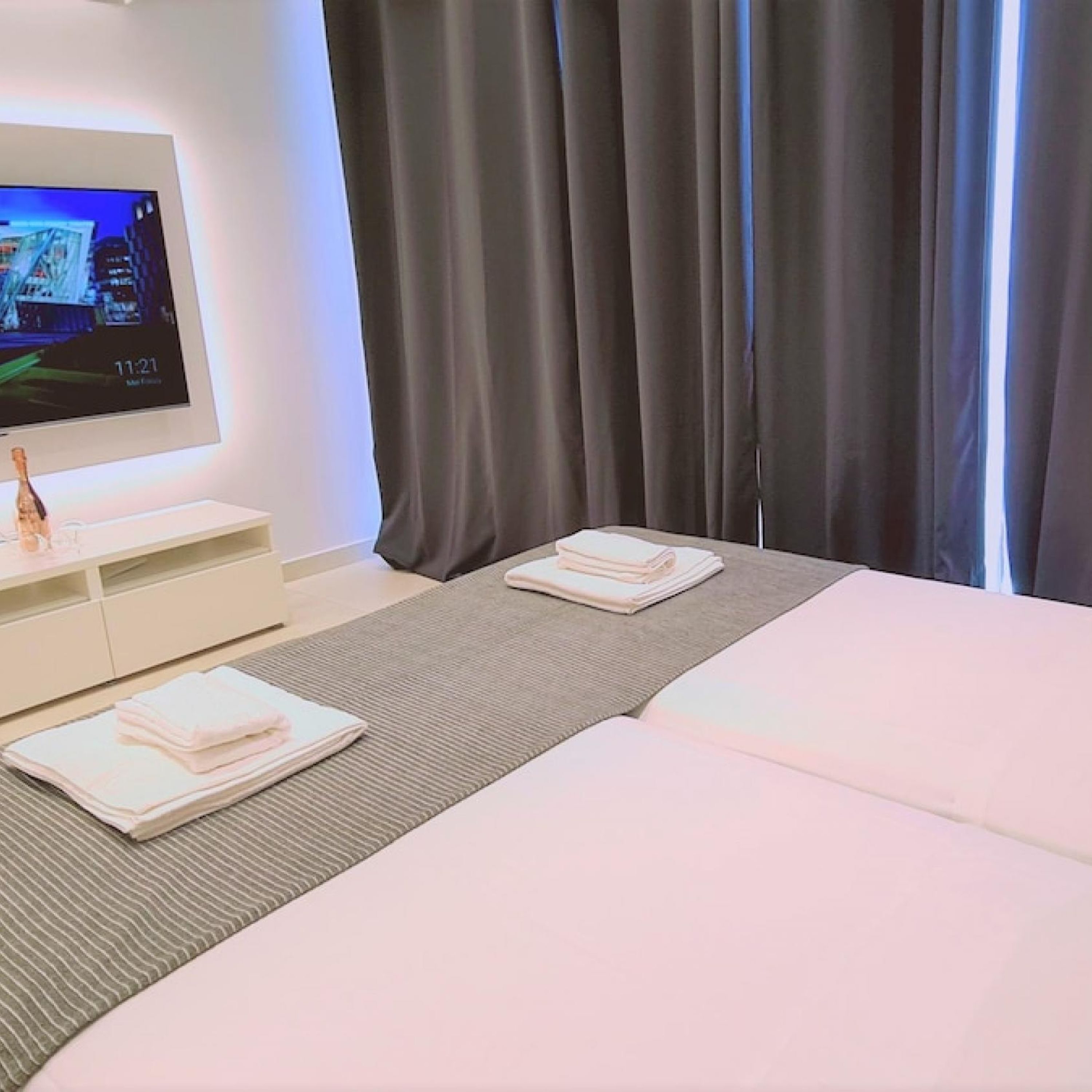 A hotel room with a bed, a TV on a stand, a small table with a bottle, and a window with curtains.