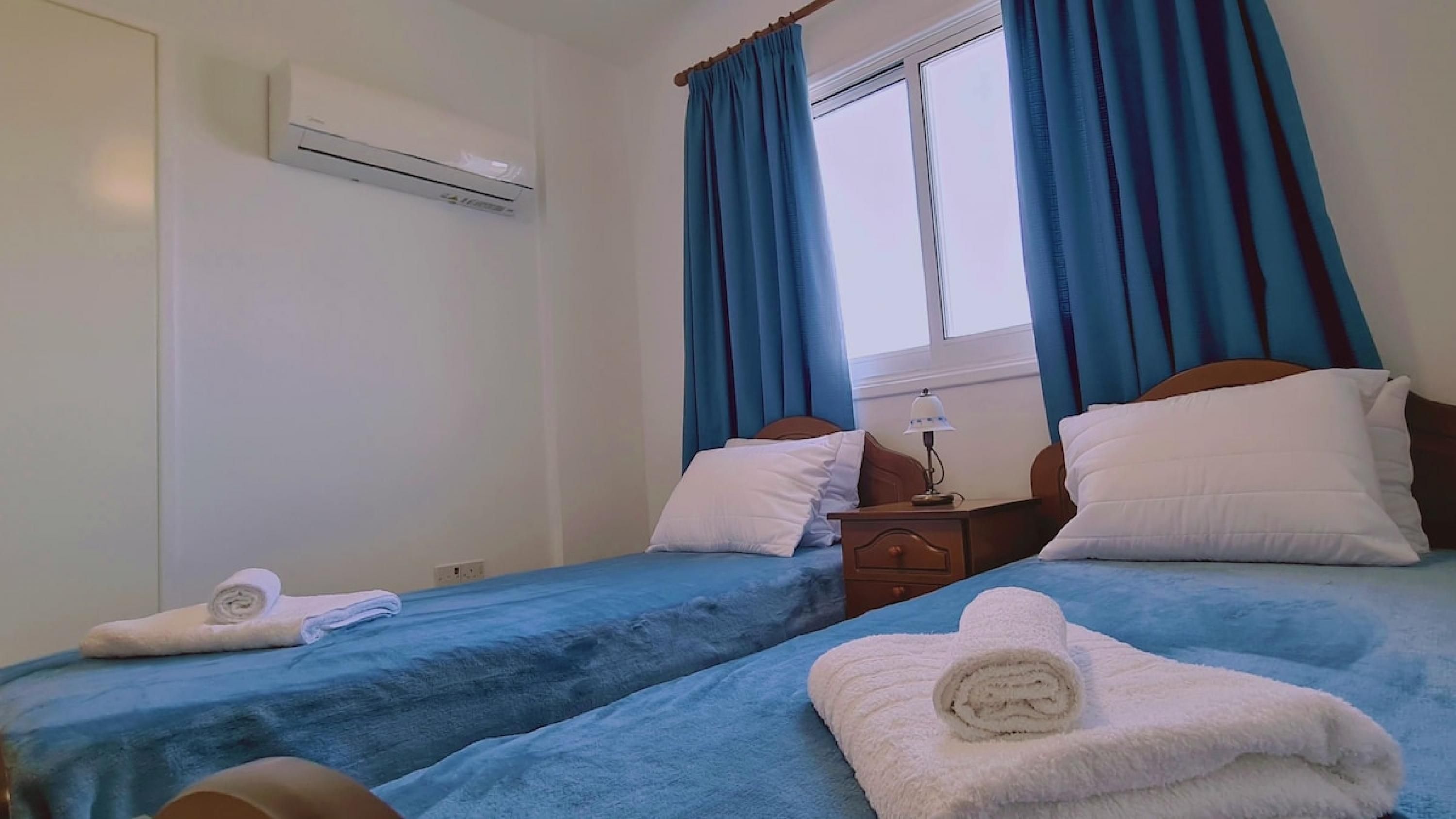 A hotel room with two beds, a nightstand, an air conditioning unit, and a window with blue curtains.