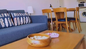 A living room with a sofa, a dining table with chairs, a kitchen area with a washing machine, and a wall-mounted air conditioner. - 35 LCA - Camelia Apartment (Larnaca)
