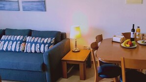 A living room with a sofa, a table with chairs, a lamp, and a dining table with fruits and a bottle. - 35 LCA - Camelia Apartment (Larnaca)