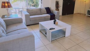 A modern living room with a sofa, a coffee table, and a dining area visible through a glass door. - 51 PER - Sunny Bay Villa (Paralimni)