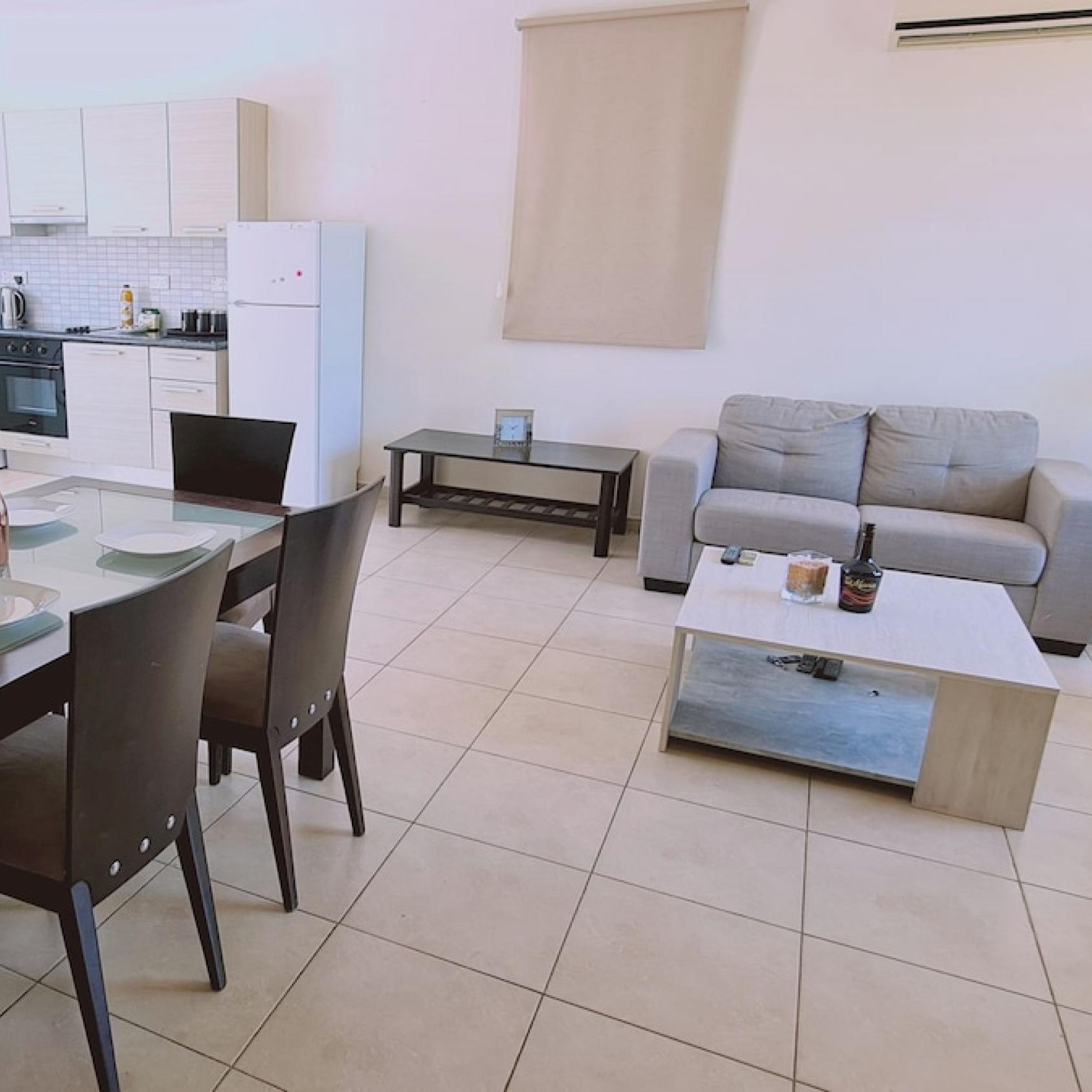 A modern kitchen with white cabinets, a white refrigerator, and a dining area with a table and chairs. There is a gray sofa and a coffee table in the living area.